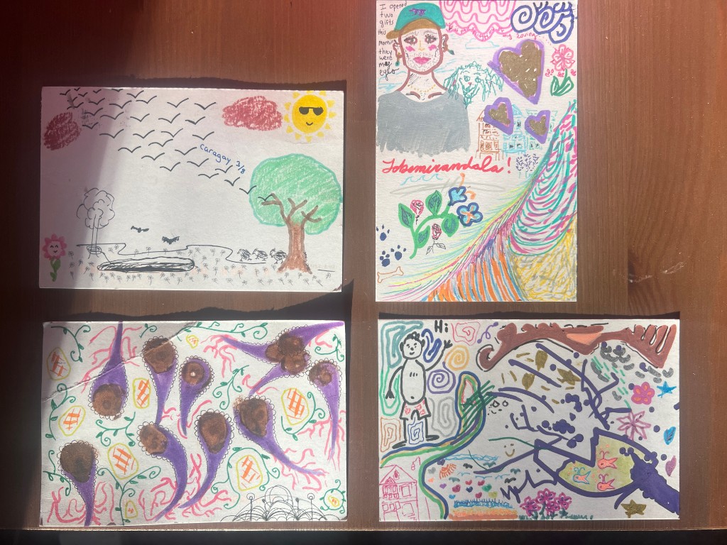 four completed postcards arranged in a grid on a wooden table, each covered in layered drawings from different hands